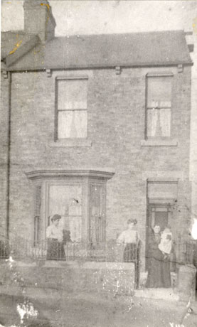 Photograph of the exterior of the front of a terraced house, with two windows on the first floor, and a bay window and door in the ground floor; in front of the bay window, behind the wall of the yard of the house, two women wearing blouses and long skirts can be seen; in the gateway, in front of the doorway, a woman in a long dress can be seen holding a baby; the photograph has been identified as A Group of The Taylor Family in Church Street, Haswell