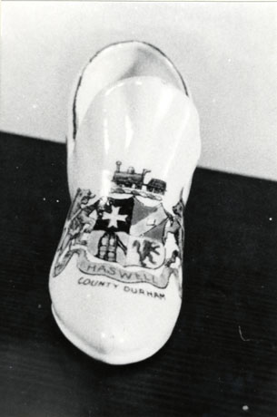 Photograph of a ceramic shoe bearing the a coat of arms and the words, Haswell County Durham; the coat of arms consists of a shield quartered with a cross of St. John, a miner's lamp, a lion rampant, and a bar sinister; it also has supporters of a lion and a bull; it is surmounted by a locomotive