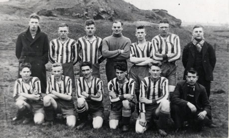 Photograph of ten members of Haswell Football Team, wearing the club strip, accompanied three other men in overcoats and scarves; the players are posed in a field with a hill behind and very faint buildings in the far distance