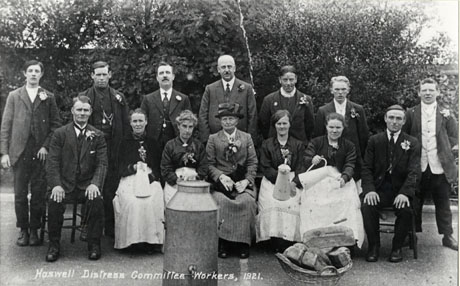 Photograph entitled Haswell Distress Committee Workers, 1921, showing a group of nine men and five posed in front of trees; the men are wearing suits and button holes in their lapels; one woman, who is also on hasw0023, is wearing a suit and button hole; the other four women are wearing long aprons and three are holding jugs; the fourth one is partly obscured by a large milk churn in front of her; also in front of the group is a whicker basket containing loaves of bread