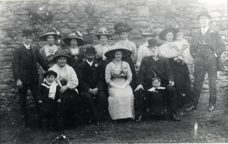 Photograph of four men and eight women and three children posed against a high stone wall; the men are in suits and have button-holes in their lapels; the women are finely dressed in elaborate dresses, blouses and hats; the photograph has been identified as A Wedding Group from Haswell