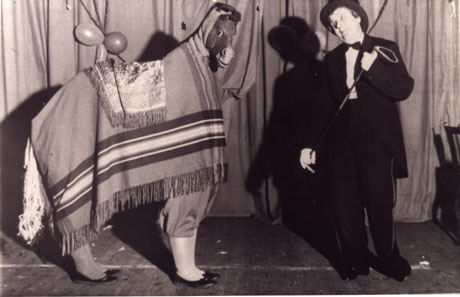 Photograph showing a woman dressed in a man's dinner jacket and trousers and bowler hat, holding a whip and standing in front of a curtain, most likely on a stage; the woman is looking ruefully at a pantomime horse, which is standing sideways to the camera; the horse has two pairs of feminine legs protruding from the blanket thrown over its back, a tail of straw is protruding from its rear and a small head may be seen at its front; the photograph has been identified as Haswell Women's Institute Pantomime