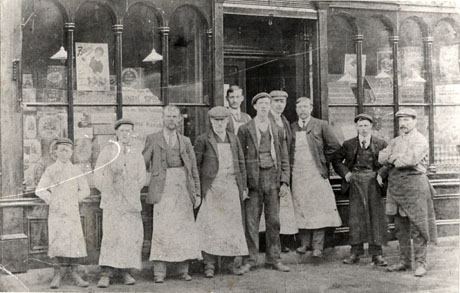 Photograph of ten men and boys outside the windows of a grocery shop; a sign advertising Bovril, tins of Co-operative Wholesale Society products and a poster advertising annual sports can be seen in the left-hand window; the contents of the right-hand window are difficult to determine; all but one of the men and boys are wearing aprons; the photograph has been identified as depicting the Haswell Co-op, 1906