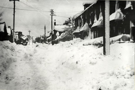 Photograph looking the length of a street, identified as Sycamore Terrace, Haswell, under heavy snow; the roofs of the houses and of the bay windows of the houses, are covered in deep snow as is the surface of the road and the gardens and pavements; telegraph poles can be seen furred with snow; the date of the photograph has been identified as 1941