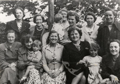 Photograph of a group of eleven women with three young children posed against a background of trees; they are young and middle-aged and the children are all under five years of age; the women are wearing coats or dresses and cardigans; the photograph is described as Haswell Garden Party