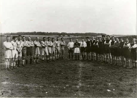 Photograph of two lines of eleven men drawn up opposite each other in a field with open countryside in the distance; between the two lines are three men standing facing the camera; the two lines of men are wearing shorts, socks and boots and would appear to be members of two football teams; the three men standing between them are also wearing shorts, socks and boots and would appear to be referee and linesmen; the photograph is entitled Haswell Football Team, Late 1940s