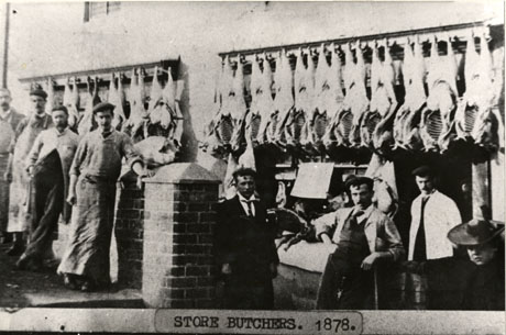 Photograph entitled Store Butchers. 1878, showing the facade of a building with the carcases of animals hanging above open butcher's shop windows; seven men, employees, it would seem, of the butcher's shop are standing in front of the two windows; at the extreme right of the photograph the head and shoulders of a woman wearing a hat can be seen