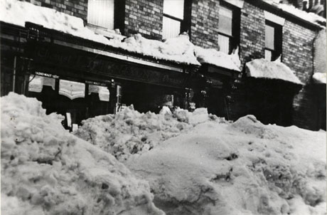 Photograph showing the windows of a shop identified as Nixon's Chemist Shop, Haswell, with snow piled against it almost to the top of the shop windows; the windows on the storey above can be seen with snow on the roofs of bay windows; the photograph has been dated as 1941