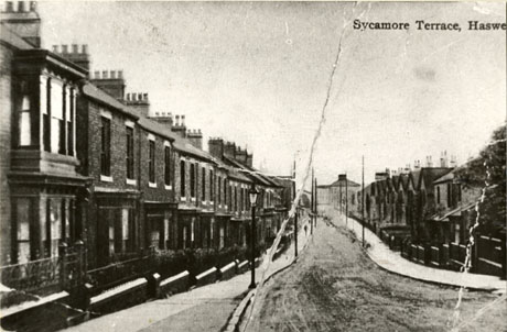 Postcard photograph entitled Sycamore Terrace, Haswell showing a street of terraced houses receding downhill from the camera; the houses on the left-hand side of the street have bay windows, small front gardens and walls topped with railings; those on most of the right-hand side have pronounced gables but no bay windows; a large building can be seen at the far end of the street