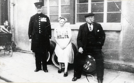 Photograph showing a man dressed as a Chelsea Pensioner; a woman wearing a dress, patterned pullover and scarf on her head; and an elderly man dressed in a suit and cap and smoking a pipe; the woman and elderly man are sitting on barrels; all three are outside a building with large windows, identified as the public house, The Oddfellows Arms; at the extreme left of the picture part of a perambulator can be seen; the elderly man has been identified as Bob Cragg; the woman as his daughter-in-law, Margaret Woods; the Chelsea Pensioner is identified as his brother-in-law