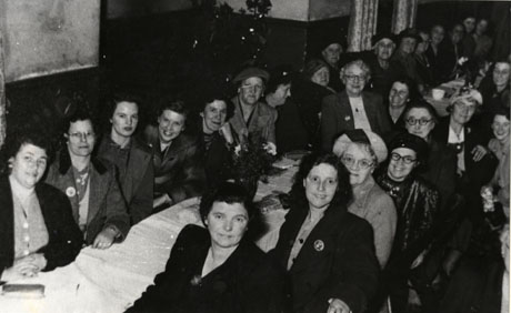 Photograph of twenty six women sitting along tables laid with white cloths in what appears to be a parish hall; all the ladies are wearing coats and hats and are described as attending Haswell Women's Institute Party, 1946