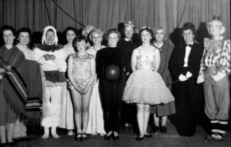 Photograph of thirteen women standing on a stage in front of curtains; they are dressed as a dancer, a clown, a wild animal, a man in evening dress, and others impossible to identify; they have been described as taking part in the Haswell Women's Institute Circus Night