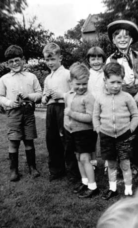 Photograph of six children, aged approximately six years, standing in a garden; three of them are wearing short trousers, another is wearing long trousers and another is dressed in cowboy clothes; two are holding a toy; they have been described as attending a garden party in Haswell