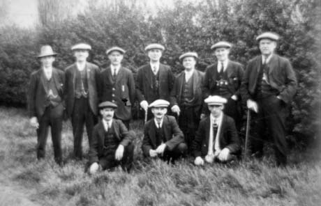 Photograph showing ten men posed in two rows in front of trees; nine are wearing caps and the tenth a hat with a high crown; all are dressed in suits and ties; three are holding walking sticks; they have been described as A Group of Haswell Gentlemen
