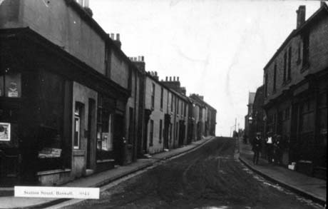 Postcard photograph entitled Station Street, Haswell., showing a narrow street running away from the camera with terraced houses on either side; the building nearest the camera on the left is a shop which cannot be identified; three indistinct men are on the right side of the street; the photograph is dark and details cannot be seen easily