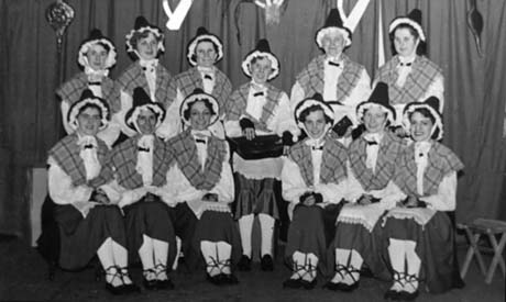 Photograph of twelve women sitting on a stage with curtains with pictures of leeks and harps on them; the women are all dressed in Welsh costume of pointed hat, checked shawl, skirt, apron, lacey blouse, and pumps; they have been identified as taking part in a Welsh Evening at Haswell Women's Institute
