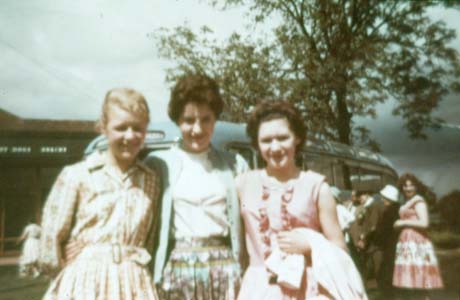 Photograph of three young women, arm in arm, dressed in summer dresses; behind them is a building, a single-decker motor bus, and two middle-aged men, two middle-aged women and a young woman, standing near the bus; the photograph ahs been described as Outing to Gretna Green from Haswell