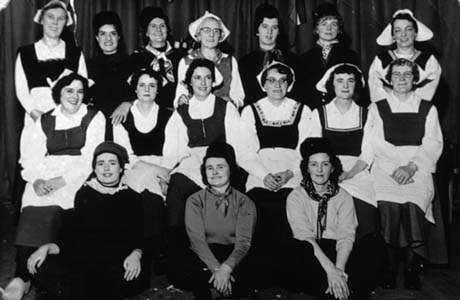 Photograph of sixteen women posed in front of curtains and dressed as Dutch men and women; they have been described as Haswell Women's Institute Go Dutch