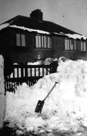 Photograph of a semi-detached house with bay windows on the ground and first floors with snow piled to the height of the garden gate in the garden and in the road outside; a spade is standing in the pile of snow outside the gate; the house has been identified as belonging to Mr. Jones of Haswell