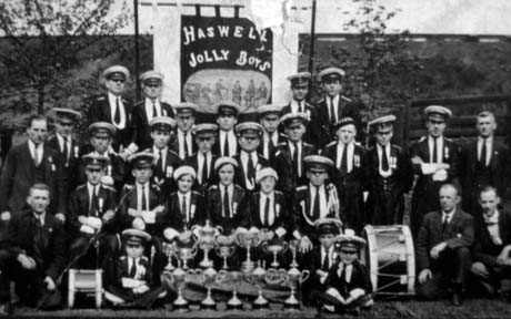Photograph of nineteen men, three women, and three small boys, aged approximately seven years, posed in uniform, accompanied by five men in suits; behind them is a banner, with the words Haswell Jolly Boys and a picture of six boys, on it; in front of the group are twelve trophy cups and two drums