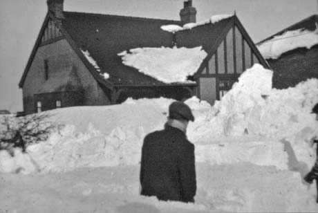 Photograph of the roof and first floor of a house with mock Tudor timbering on it, showing above a pile of snow; the back of the head and torso of a man, dressed in overcoat, scarf and cap, can be seen sticking out of the snow as he surveys the house; part of another man can be seen at the right of the photograph; the picture has been identified as being in Haswell