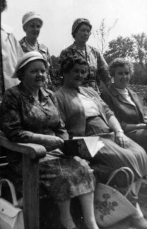 Photograph of three middle-aged women sitting, and three middle-aged women standing behind them; there are trees in the background and the women are wearing dresses and skirts and cardigans; they have been identified as members of Haswell Women's Institute on an outing