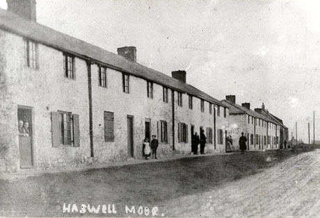 Photograph showing two blocks of houses, described as Aged Miners' Homes at Haswell Moor; in the block closer to the camera six separate dwellings can be identified; each dwelling consists of one door, one window on the ground floor and two windows on the first floor; seven indistinct figures can be seen in front of the buildings and two indistinct figures are looking over the door of one of the dwellings