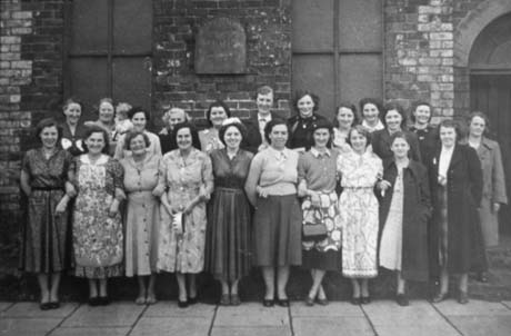 Photograph of twenty two women dressed in frocks, skirts, and overcoats, standing posed in two rows on the pavement outside a brick building, which may be a chapel; they have been described as Coronation Party at Front Street, Haswell