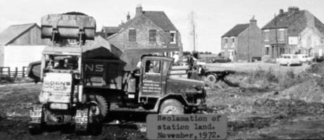 Photograph with a typed note on it as follows: Reclamation of Station Land. (Haswell) November, 1972, showing open ground with houses in the background and the side of a lorry in to which a vehicle with caterpillar tracks is putting soil; behind the lorry more machinery and two men can be seen indistinctly This photograph was submitted by Philip Soakell from the album 'History of Haswell and District', which was compiled by his grandfather Fred Soakell.