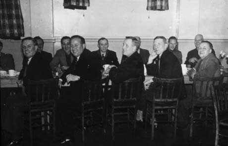 Photograph showing ten men and one woman sitting either side of a table on which cups are standing; a painted wall and window curtains are behind them; the men are all wearing suits; they have been identified as being in I.C.I. Canteen, Haswell