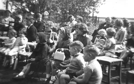 Photograph of thirteen children, aged approximately between two and six years, sitting in rows with trees behind them; they are accompanied by approximately six women; some of the children are wearing raincoats; they appear to be looking at something out of shot to the left; they have been identified as attending a Garden Party at the Manor House, Haswell