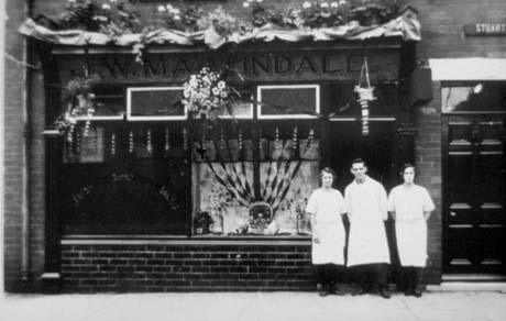 Photograph of a man and two women, wearing overalls and aprons, standing outside a shop with flowers above the window and a basket of flowers and two decorations in front of it; there are streamers in the window, and the door to the shop is closed; it is not possible to determine the occasion on which the photograph was taken; the name Martindale can be seen above the shop and the words Fresh Supply Daily can be seen on the window; the shop has been identified as being a fish shop in Haswell; the word Stuart can be seen above the door to the shop