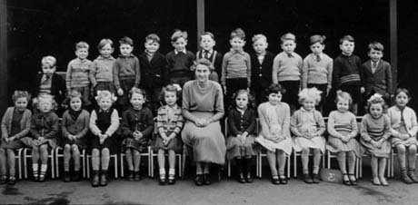 Photograph of twenty five children, aged approximately five years, posed in two rows, with a woman sitting on the front row; she has been identified as Miss Whitfield and the children as members of a class at an infant school in Haswell