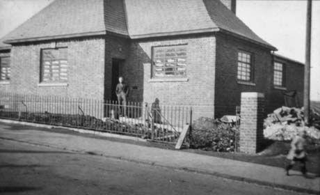Photograph showing an indistinct figure of a man standing outside the entrance to a single-storey brick building with large reinforced windows; there appears to be rubble in the grounds of the building, which has been identified as the Dole Office in Haswell; the man has been identified as Jack Whitfield