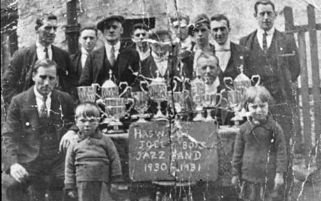 Photograph showing nine men standing behind, and two men sitting at, a table on which there are ten trophy cups; a small boy and a small girl, aged approximately six years, are standing in front of the table; in front of the table is a notice reading: Haswell Jolly Boys Jazz Band 1930-1931