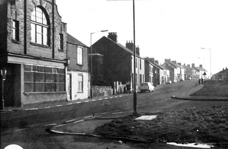 Photograph showing one side of a street running uphill from left to right of the picture; a large building is at the bottom of the slope followed by houses; a van and two cars are parked in the road; two open spaces can be seen on the opposite side of the road, which has been identified as Front street, Haswell This photograph was submitted by Philip Soakell from the album 'History of Haswell and District', which was compiled by his grandfather Fred Soakell.