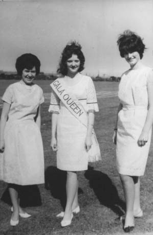 Photograph of three young women standing in a field wearing light-coloured dresses and light court shoes; one in the middle is wearing a sash reading: Gala Queen; she has been identified as being Gala Queen in Haswell