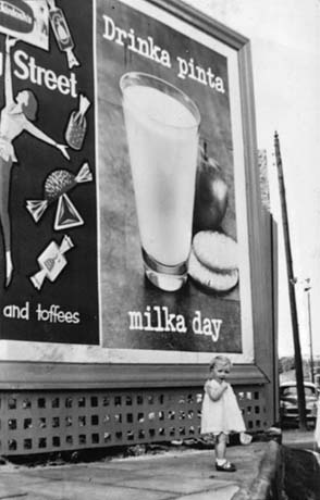 Photograph of a small girl, aged approximately two years, standing on the pavement in front of a billboard advertising milk; the advertisement has the slogan Drinka pinta milka day and shows a picture of a glass of milk, two biscuits and an apple; part of an advertisement for Quality Street confectionery is next to the advertisement for milk; the photograph has been described as Child at Corner of Co-Op, Haswell