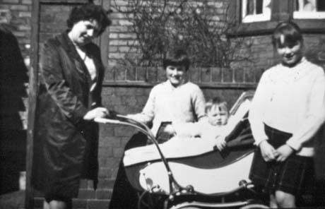 Photograph of a woman wearing a coat standing with her hands on the handle of a perambulator, in which there is an infant, whose face can be seen; a girl, aged approximately nine years, wearing a cardigan, is standing behind the perambulator and a girl, aged approximately twelve years, wearing a skirt and cardigan, is standing at the end of the perambulator; they have been identified as Mrs. A. Williams and Family