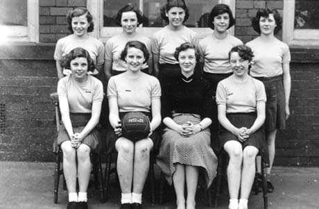 Photograph of eight girls, aged approximately fifteen years, posed in two rows in front of a brick building; a woman in jumper and skirt is posed with them on the front row; a girl on the front row is carrying a ball on which the date 1952-53 is written; they have been identified as a school netball team in Haswell