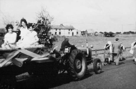 Photograph showing a tractor, registration LML 235, towing a float, on which three girls in light dresses can be seen sitting; in front of the tractor the backs of six people walking along the road can be seen; beyond the road is a field across which a low light coloured building, presumably a farmhouse, can be seen; the photograph has been described as Carnival Day, Haswell