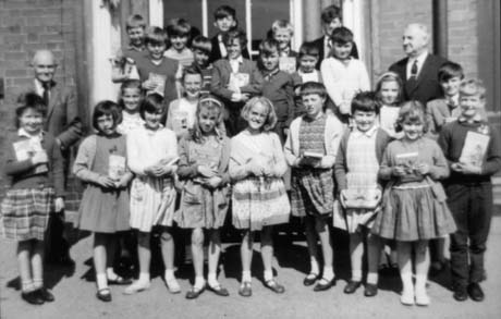 Photograph of twenty two children, aged approximately nine years, posed in the doorway of a brick building; each child appears to be holding a book; a young man, presumably a teacher, is standing at the back of the group; a middle-aged man is standing either side of the group; these have been identified as Mr. Hall and Mr. Attlee at Haswell School