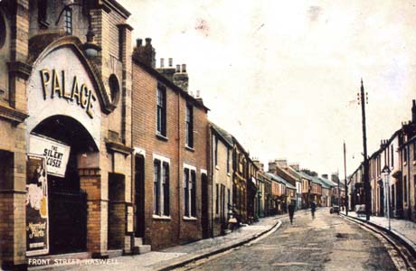 Postcard photograph entitled Front Street, Haswell, showing a street of houses and shops running away from the camera; a car is parked in the distance and indistinct figures are walking in the road and on the pavement; in the foreground on the left is the facade of the Palace cinema; it is built of bands of coloured brick and is advertising The Silent Accuser and Married Flirts