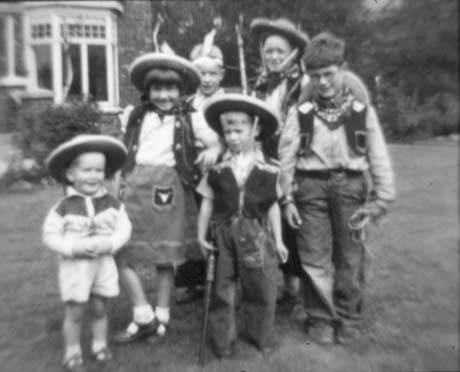 Photograph of five boys and one girl, aged between two and nine years, wearing cowboy outfits, standing on grass with a house with a bay window in the distance behind them; they have been described as Cowboys and Indians at the Manor House, Haswell