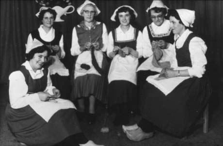 Photograph of six women dressed in dark skirts, dark bodices, and light-coloured caps, as Dutch women, sitting knitting; they are possibly members of Haswell Women's Institute and have been described as Dutch Night Singing Knitting Song