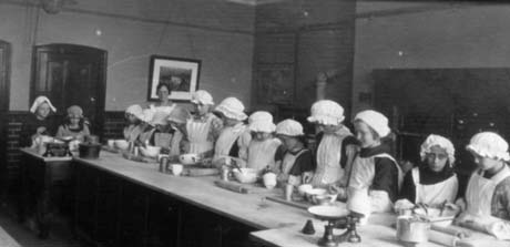 Photograph of fourteen girls, aged approximately twelve years, standing on one side of a long table, on which there are bowls, rolling pins, scales, and saucepans; the girls are dressed in aprons and caps; a woman in an apron is standing behind the girls; the walls of the room are painted in a dark colour with one picture on them; the photograph has been identified as a Cookery Class in Haswell