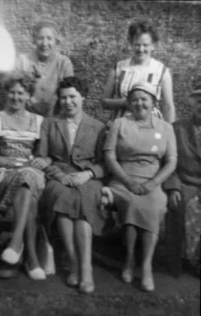 Photograph of three women sitting and two women standing behind them; there appears to be a wall behind them; they are dressed in summer frocks and have been described as members of Haswell Women's Institute on an outing