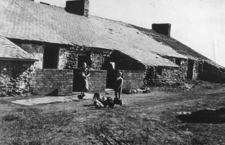 Photograph showing the exterior of single-storey cottages built of rough stone; the roof of the cottage furthest away from the camera has lost its slates and is in ruins; a woman and a boy are standing in front of one cottage feeding hens; the cottages have been identified as Low Row, Haswell This photograph was submitted by Philip Soakell from the album 'History of Haswell and District', which was compiled by his grandfather Fred Soakell.