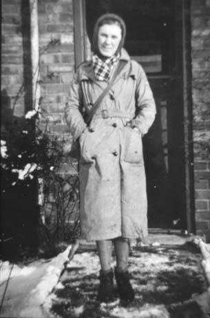 Photograph of a woman wearing a mackintosh, a hat, a scarf, and boots, and carrying a bag over her left shoulder, standing in snow in front of the doorway of a house; she has been identified as Renee Wood, the Post Woman