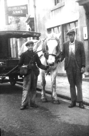 Photograph showing a boy, aged approximately fourteen years and a man standing either side of the head of a light-coloured horse harnessed to a milk float; the boy is wearing a cap and a jacket and is carrying a leather satchel over his left shoulder; the man is dressed in a cap, jacket and jacket under the outer one; behind them is a building bearing a sign reading: Oddfellows Arms; they have been identified as being Co-Op Milkman in Haswell
